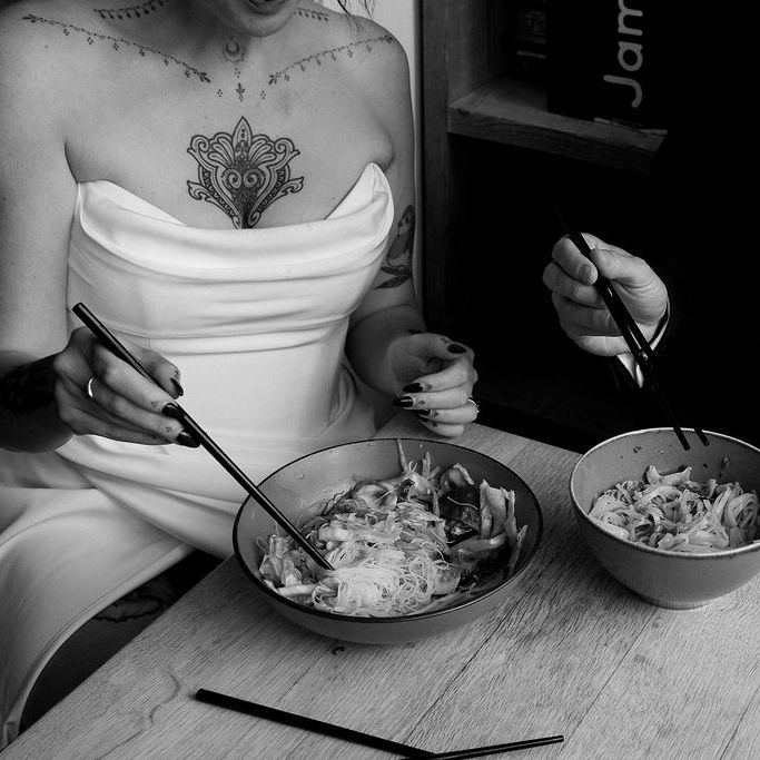 Bride and groom eating noodle bowls with chopsticks at a wooden table in black and white