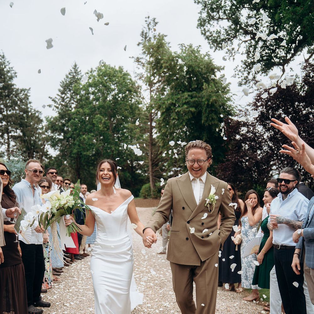 Bride and groom walk hand in hand through a petal confetti shower lined by cheering guests