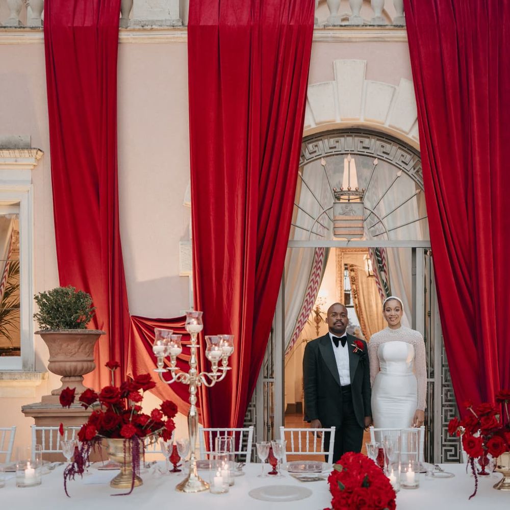 Bride and groom stand in an archway framed by red drapery above a table of red roses and candles