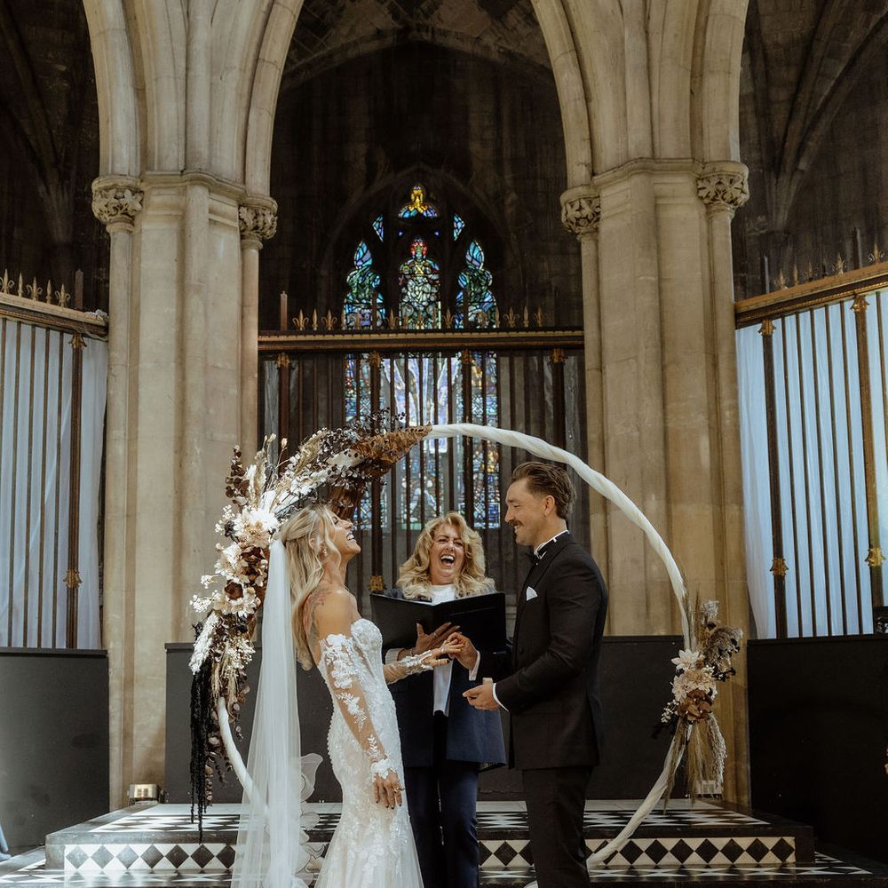 Bride and groom exchange rings beneath a floral moon arch during a Gothic chapel ceremony