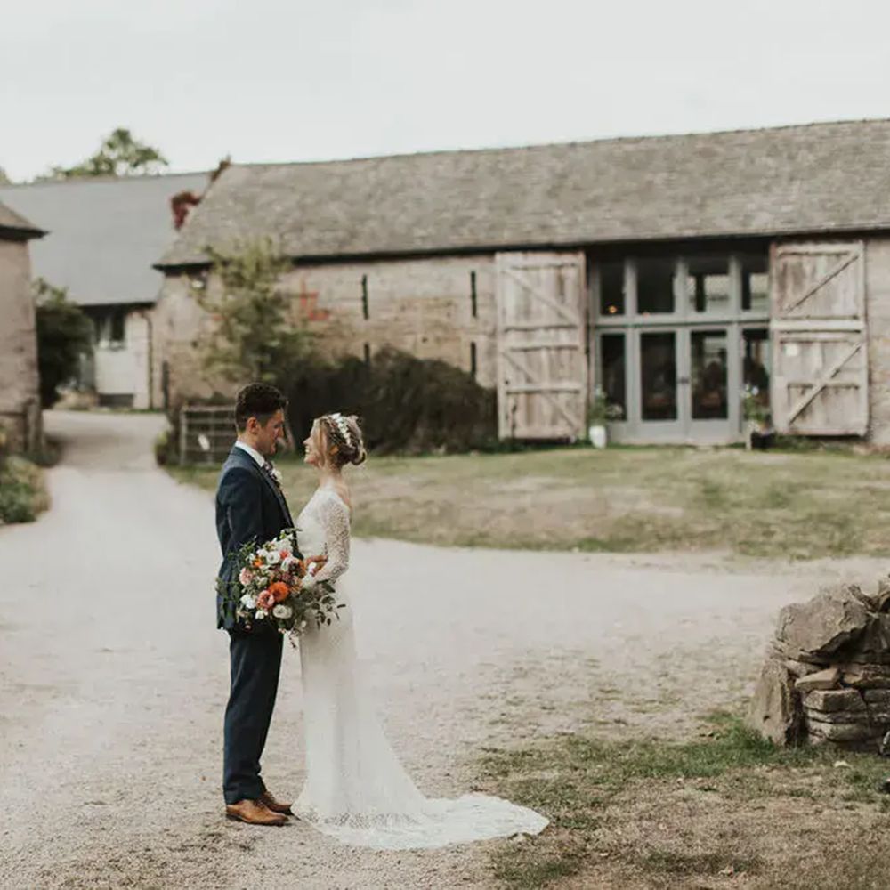 Bride and groom stand face to face in a rustic stone barn courtyard holding a bouquet