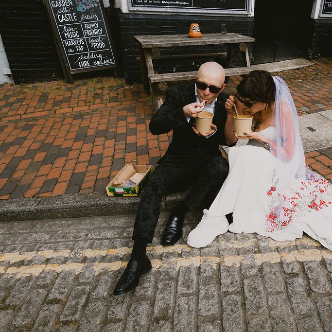 Bride and groom in sunglasses eating street food from cartons on a kerb outside a pub