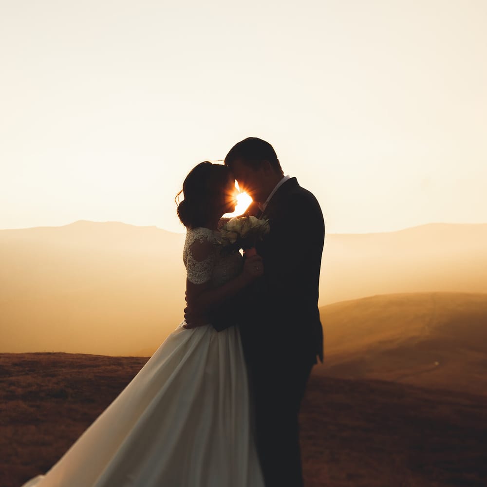 Bride and groom embrace at golden hour against a mountain backdrop with the sun between them