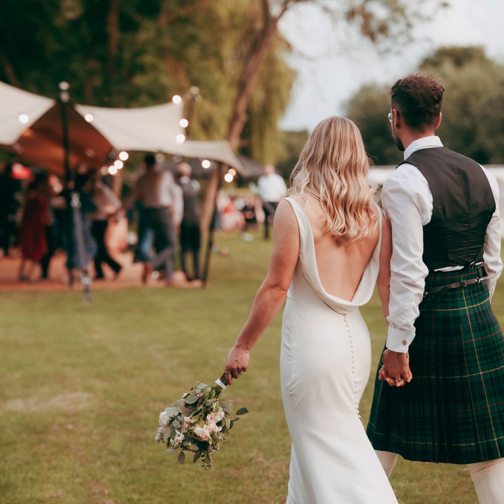 Bride in a cowl-back gown and groom in a green tartan kilt walk hand in hand towards a stretch tent