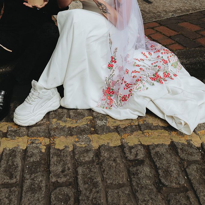 Bride in floral-embroidered gown and chunky trainers sits with groom in black on cobbles