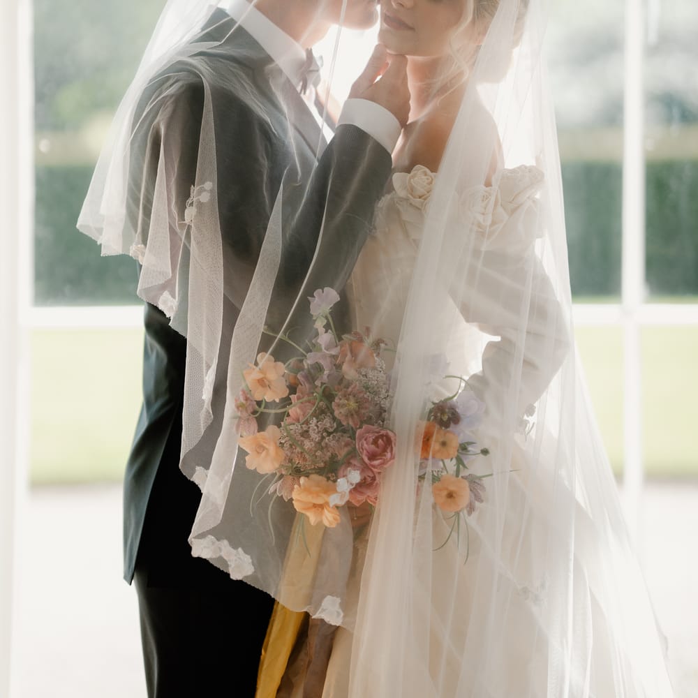 Bride and groom embrace beneath a floral-edged veil holding a pastel bouquet by tall windows