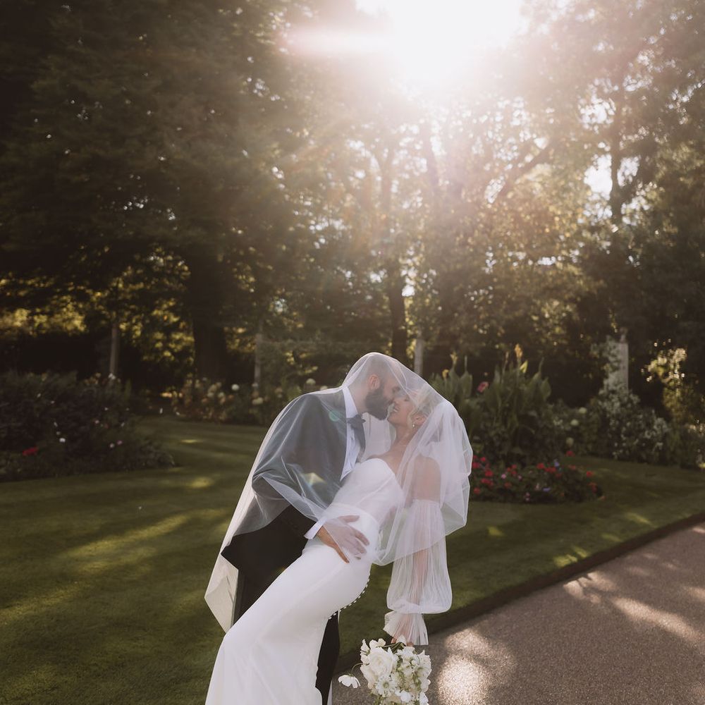 Bride and groom kissing beneath a veil on a garden path in golden sunlight