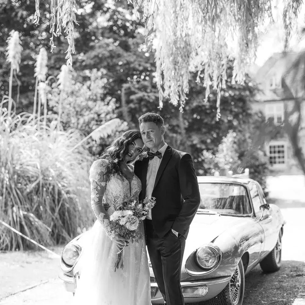 Bride and groom leaning together beside a vintage convertible car under a willow tree