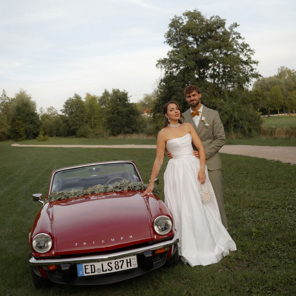 Bride and groom standing beside a vintage red Triumph convertible in a green field