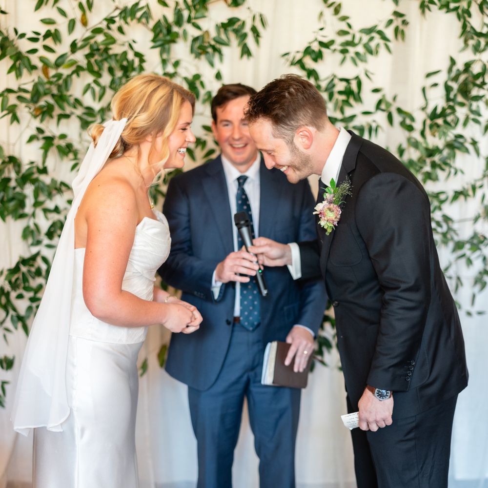 Laughing bride and groom exchange vows before an officiant beneath a foliage backdrop