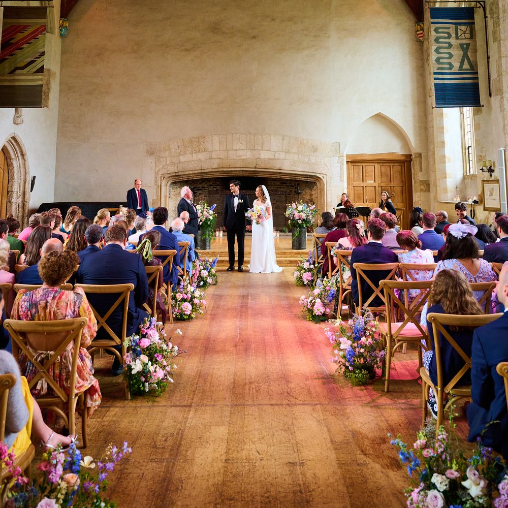 Bride and groom exchange vows before guests in a beamed medieval hall lined with wildflowers