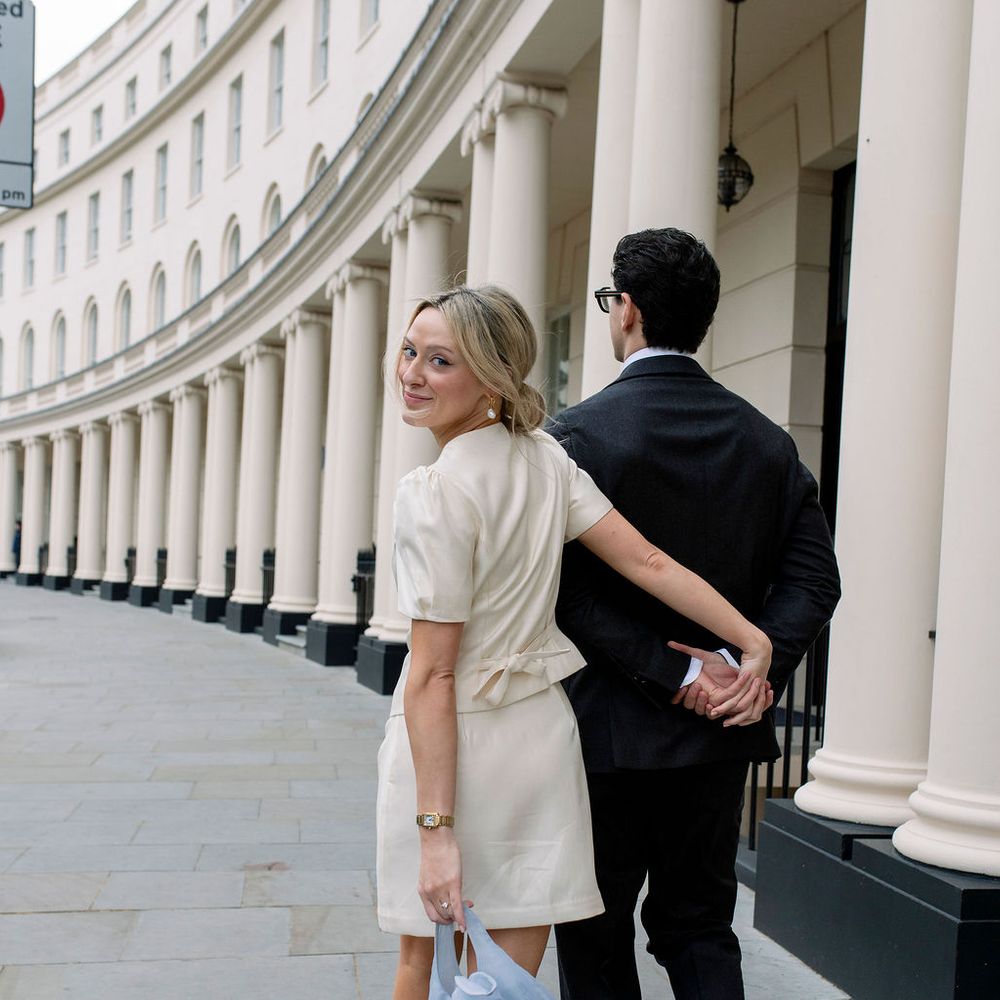 Bride in short ivory dress glances back while walking arm-in-arm with groom past white columns