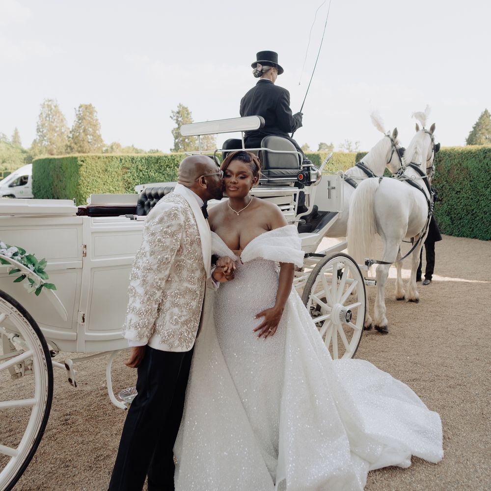 Groom kissing bride's cheek beside a white horse-drawn carriage with a top-hatted coachman
