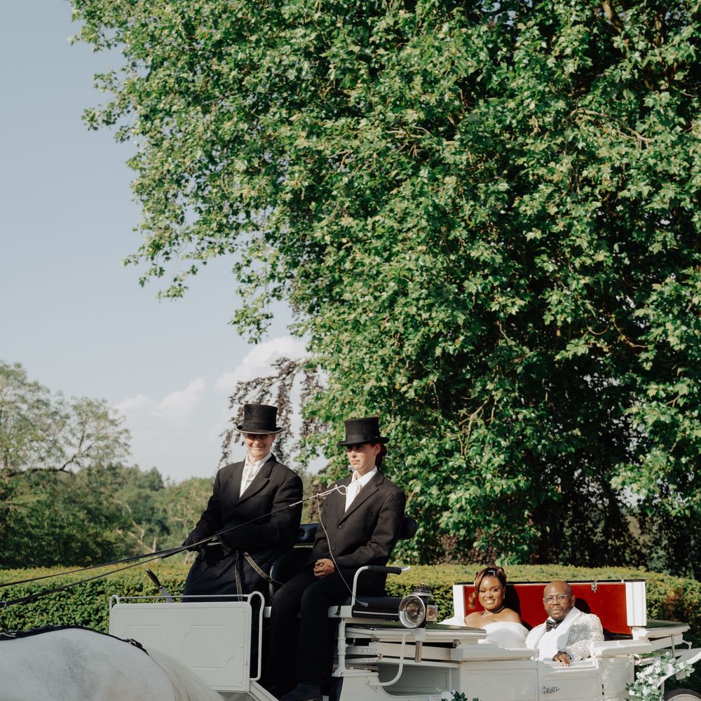 Newlyweds ride in a white horse-drawn carriage with two top-hatted drivers past leafy greenery