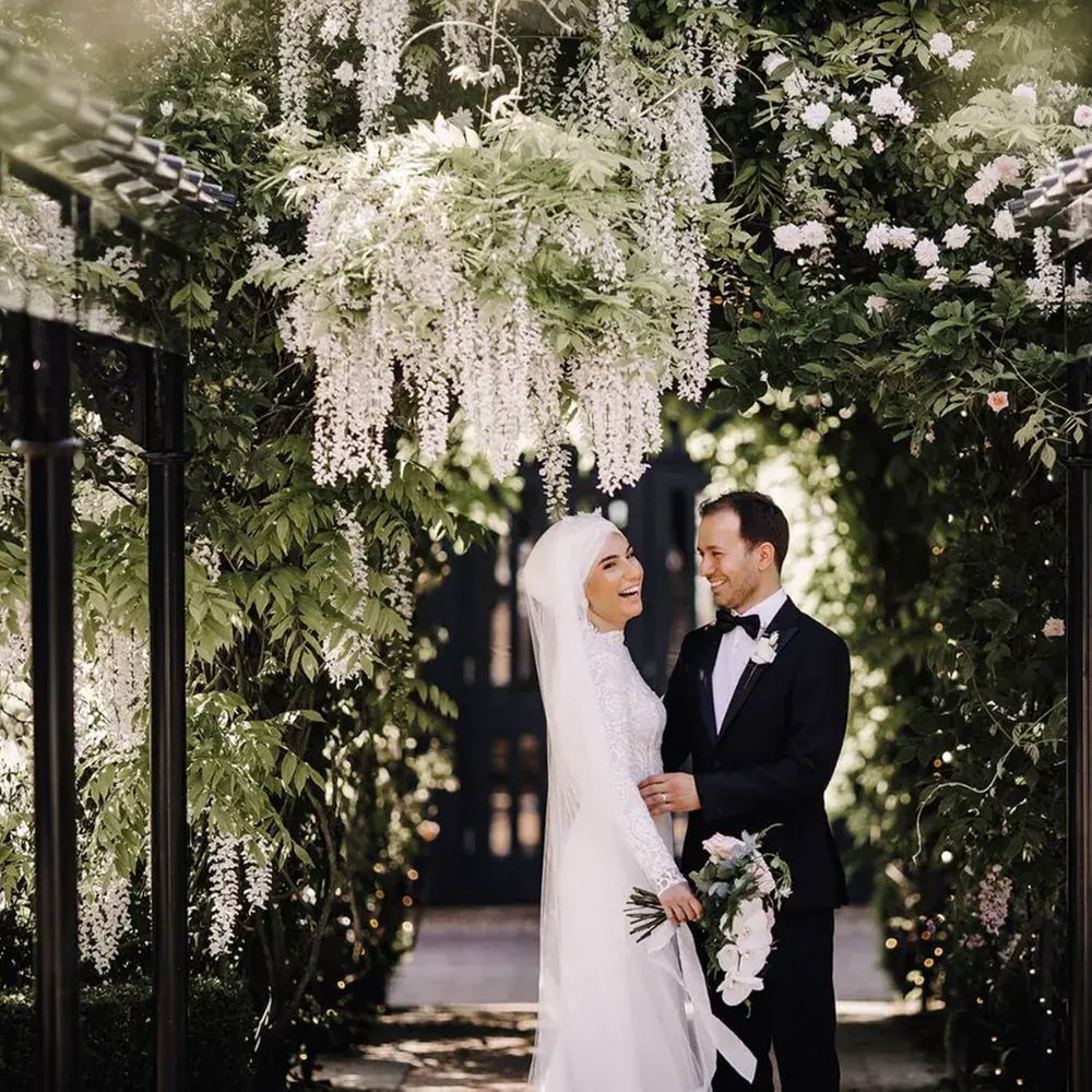 Smiling bride and groom beneath a wisteria-draped pergola in a lush garden setting