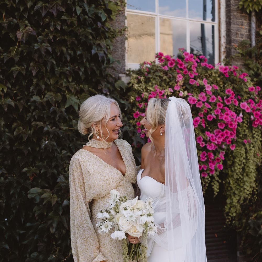 Bride in strapless gown and veil shares a smile with a guest holding a white bouquet by an ivy-clad wall