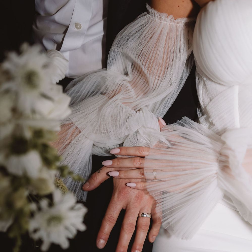 Bride's hands with wedding rings resting on a sheer tulle sleeved gown beside a white bouquet