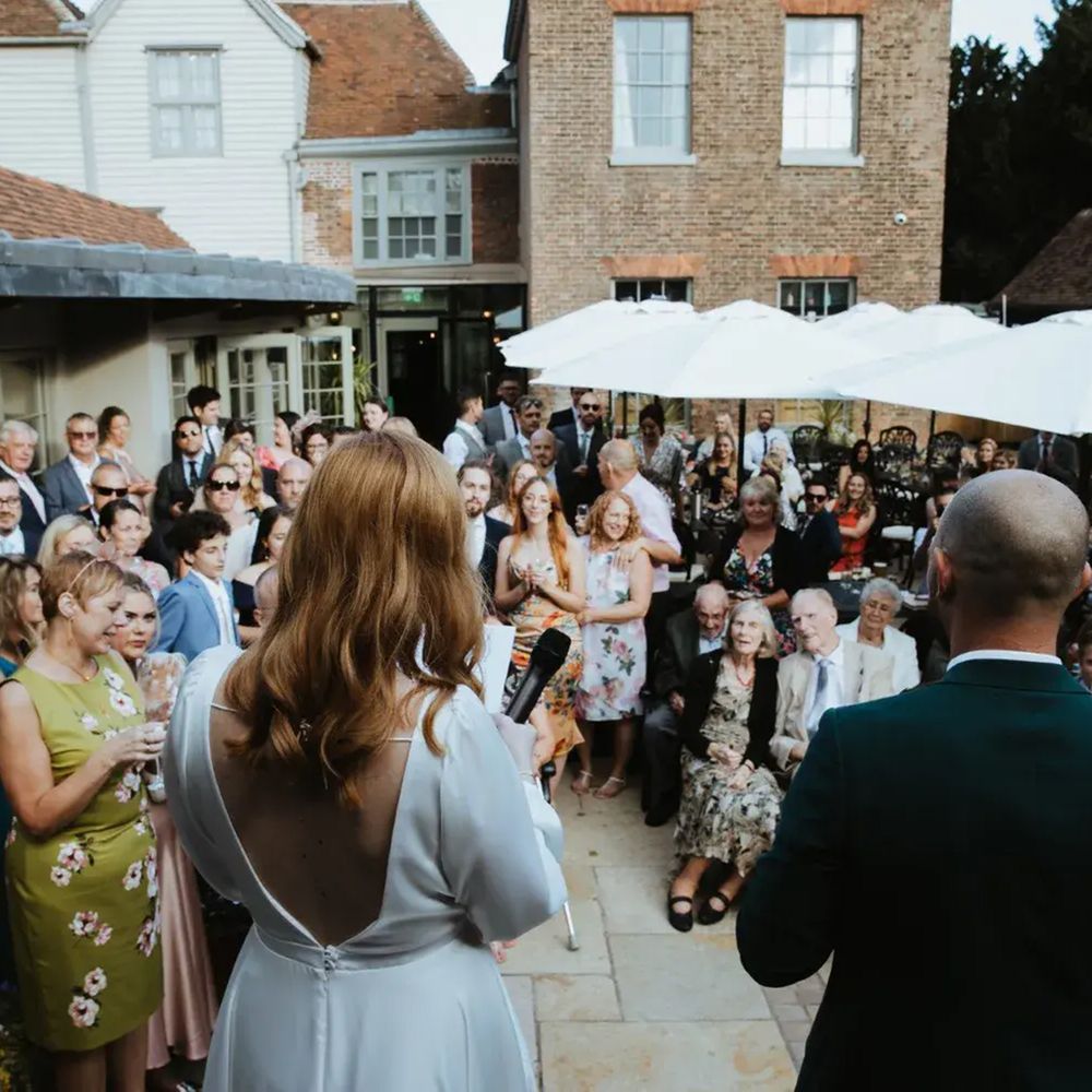 Bride holding a microphone addresses seated guests during an outdoor courtyard speech