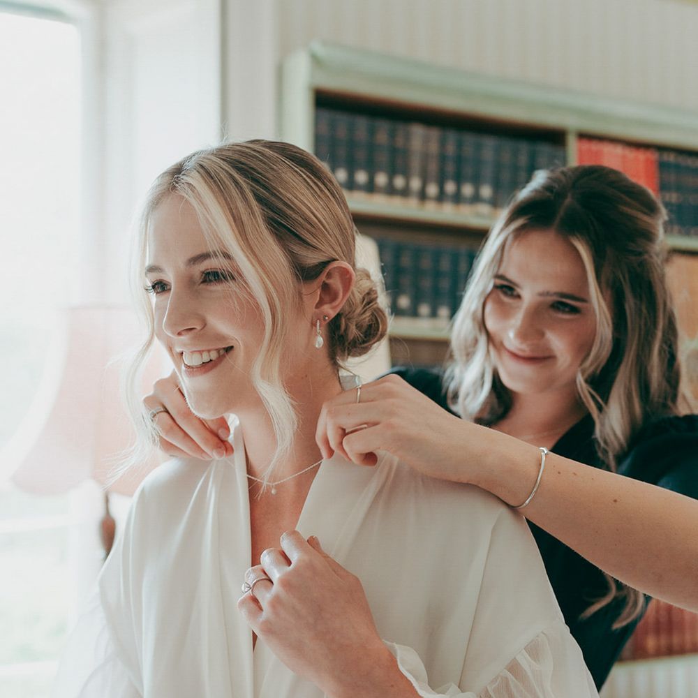 Smiling bride in a white ruffled robe having her necklace fastened during morning preparations