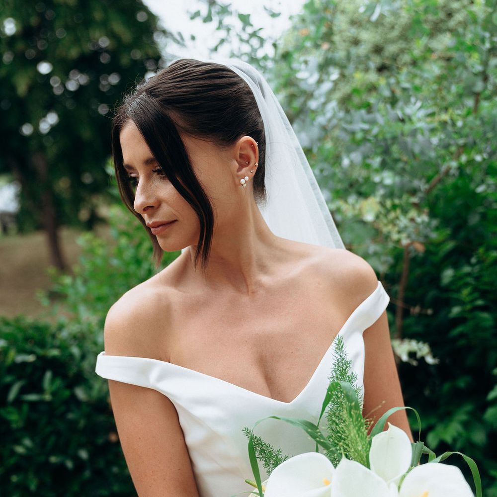 Bride in off-shoulder gown holding a white calla lily and orchid bouquet in a garden