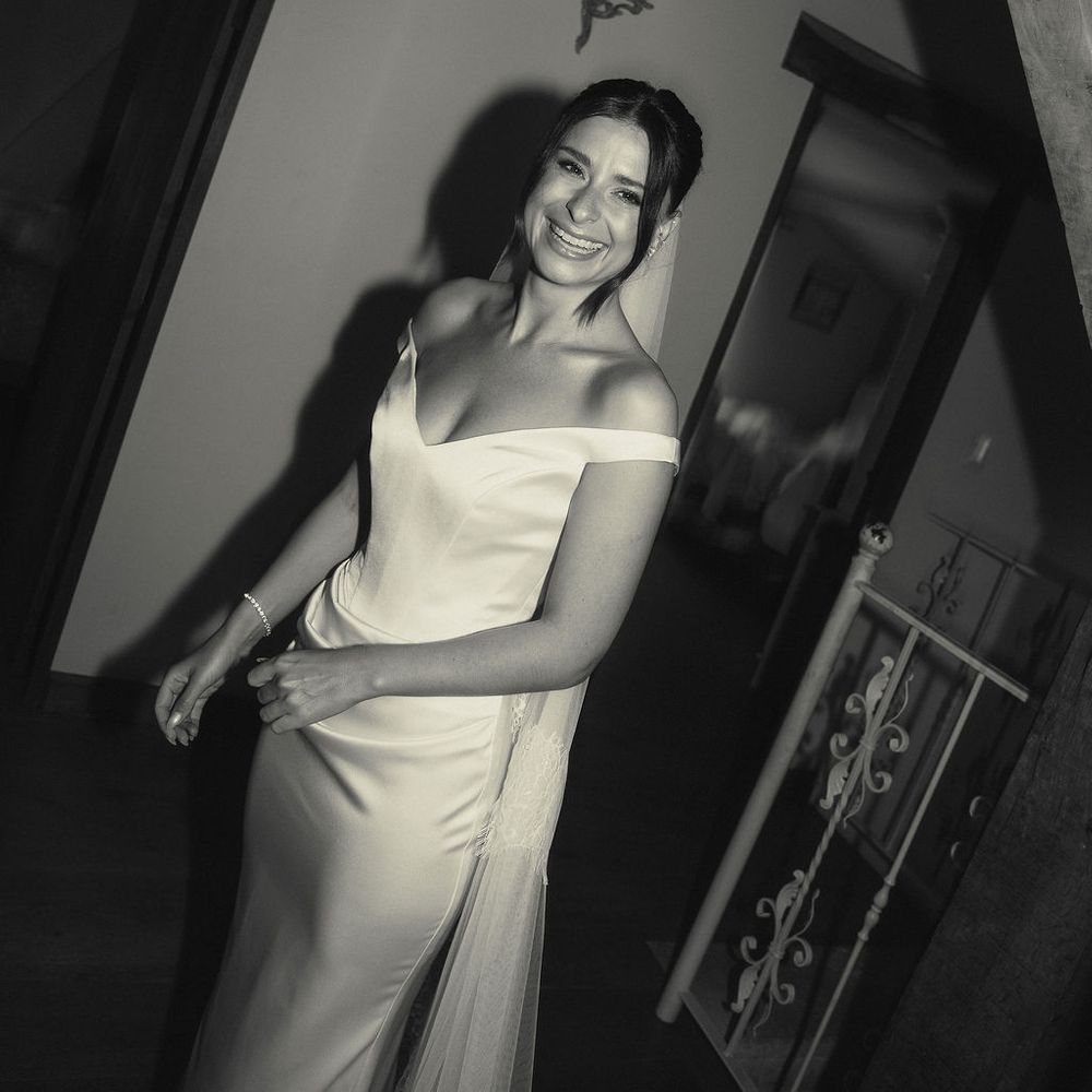 Smiling bride in an off-the-shoulder satin gown beside a wall light on a landing