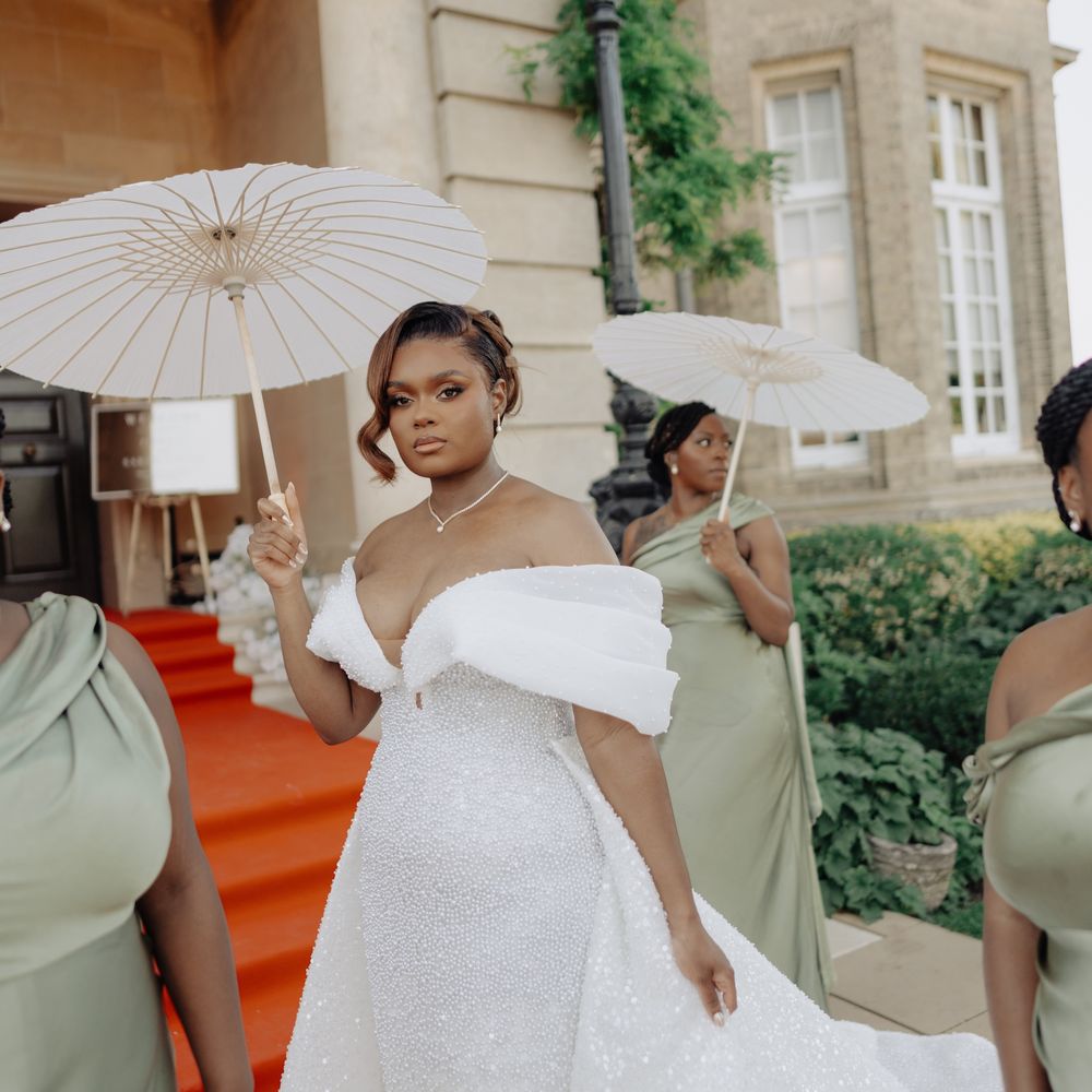Bride in a beaded off-shoulder gown holding a white parasol beside bridesmaids in sage green