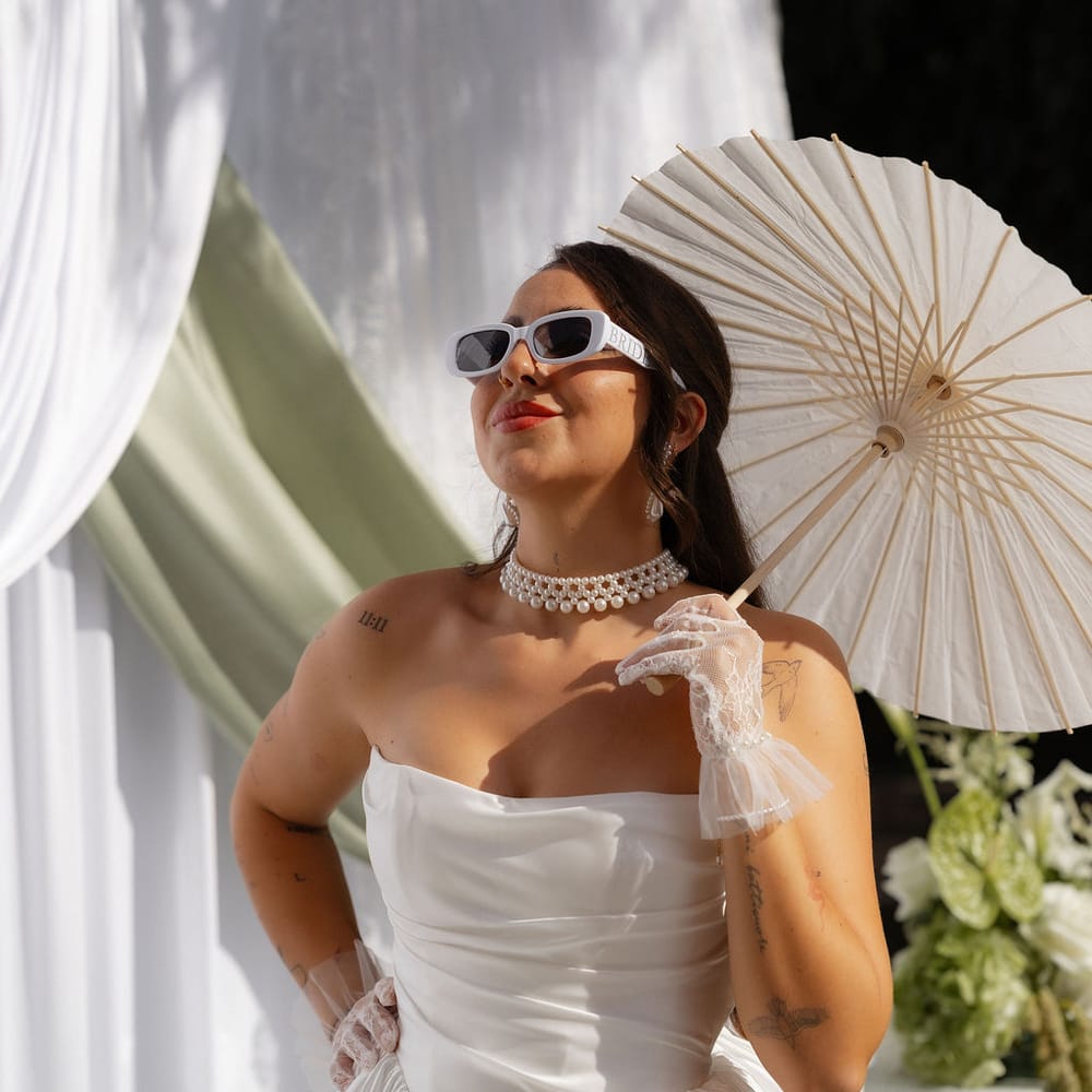 Bride in strapless gown and white sunglasses holds a paper parasol beside draped backdrop