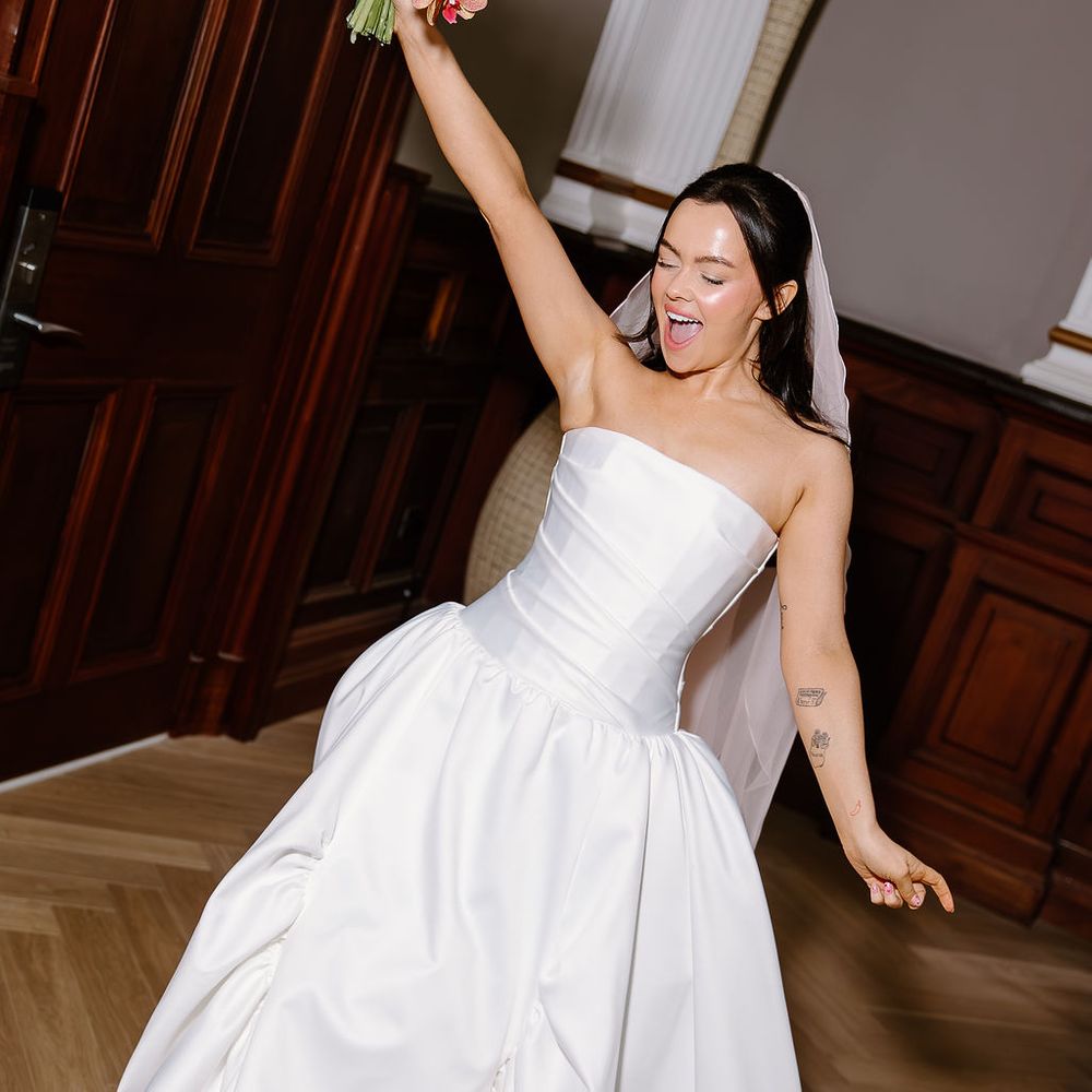 Joyful bride in strapless gown raises pink orchid bouquet aloft, wearing green heels
