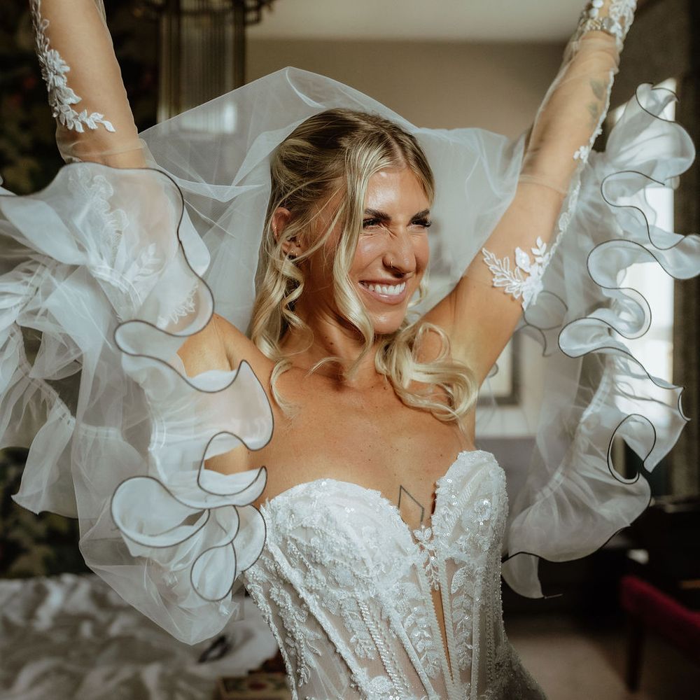 Smiling bride raises her arms holding a ruffled tulle veil in a bright dressing room