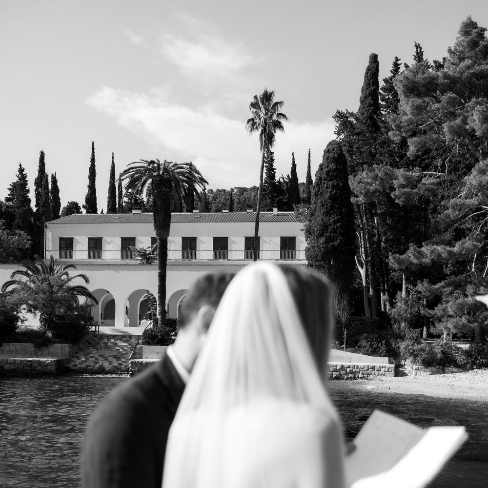 Bride in flowing veil reads her vows beside groom facing a Mediterranean waterside villa