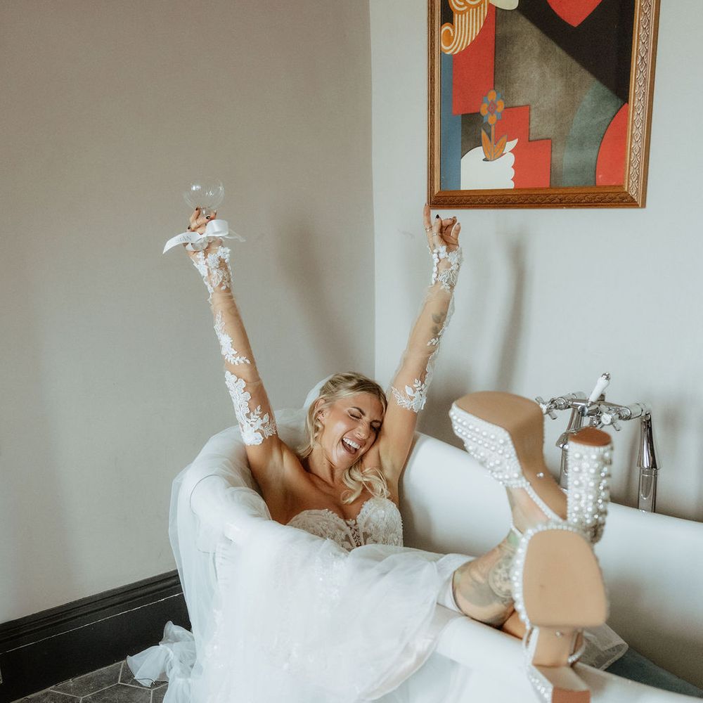 Laughing bride reclining in a freestanding bath, arms raised holding a coupe glass, pearl heels on show