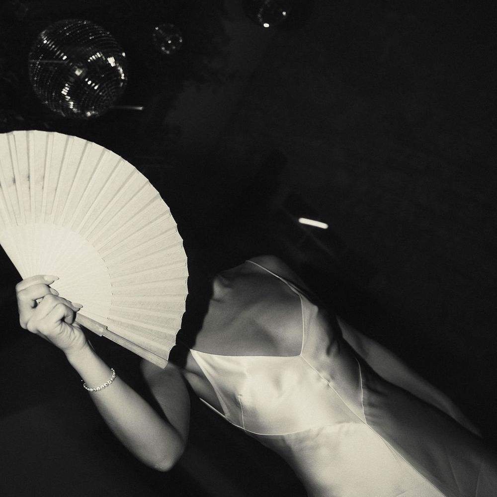 Bride in a satin slip dress holding a paper fan beside a disco ball in moody light