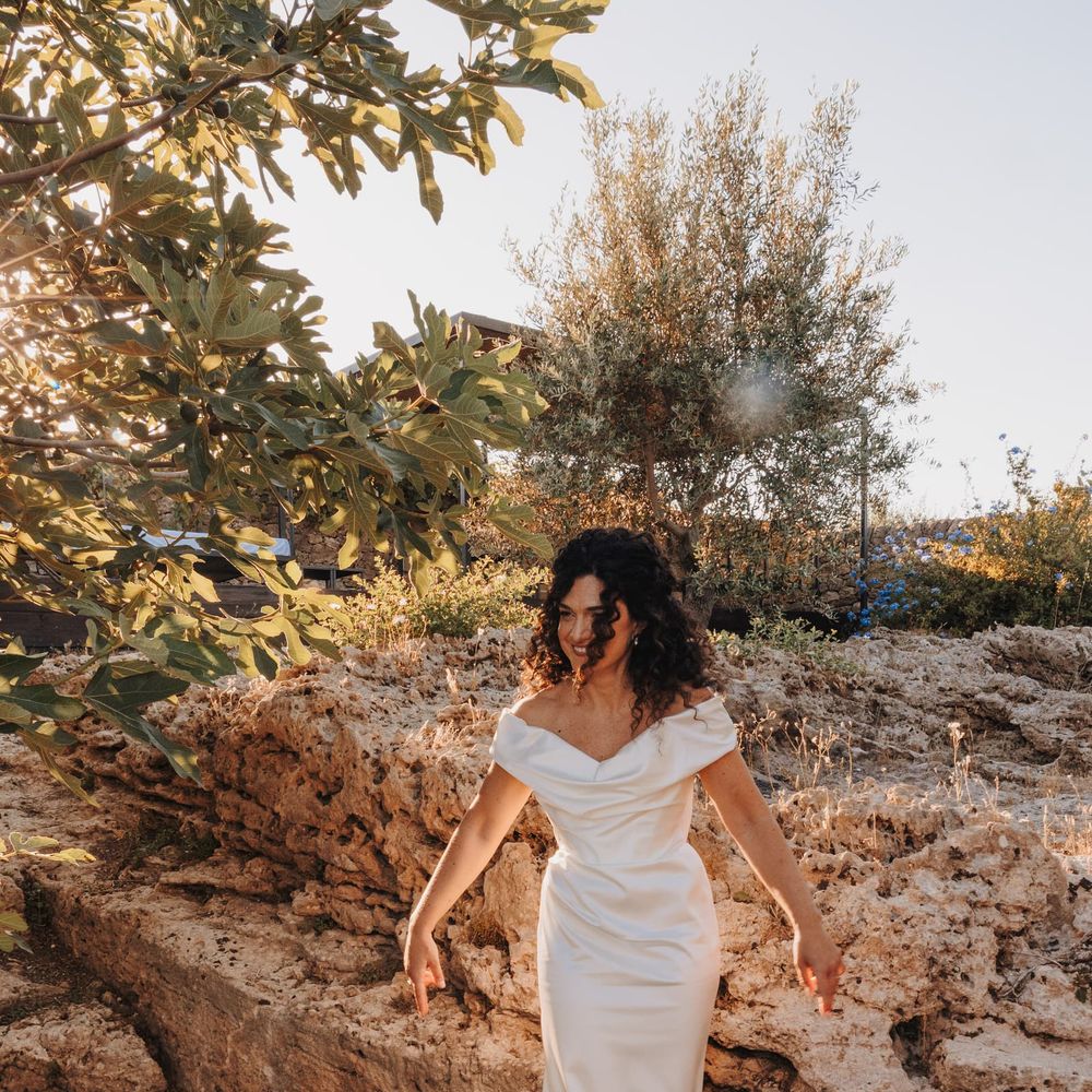 Bride in an off-the-shoulder satin gown standing among sunlit rocks and olive trees