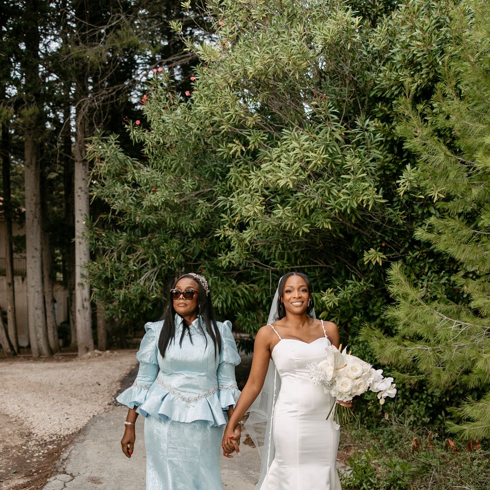 Smiling bride in a satin gown holds hands with a guest in pale blue amid lush greenery