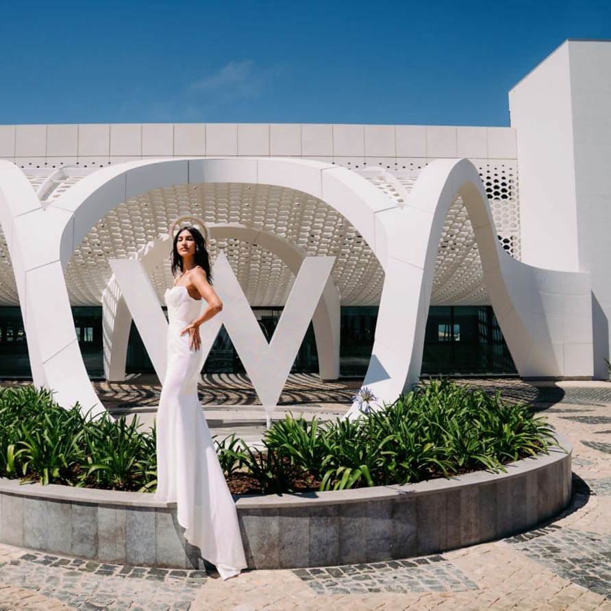 Bride in a sleek fitted gown poses before a sculptural white archway under a clear blue sky