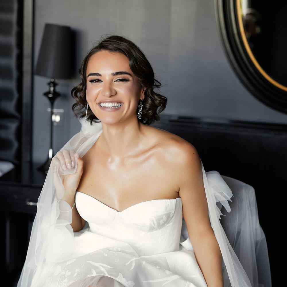Smiling bride in a strappless short tulle gown with drop earrings seated in a dark interior