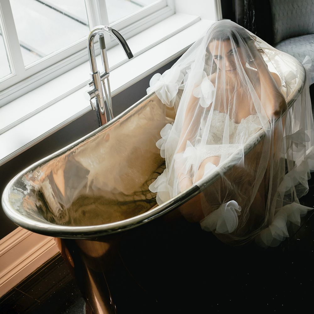Bride draped in a tulle veil sitting in a copper roll-top bath by a bright window