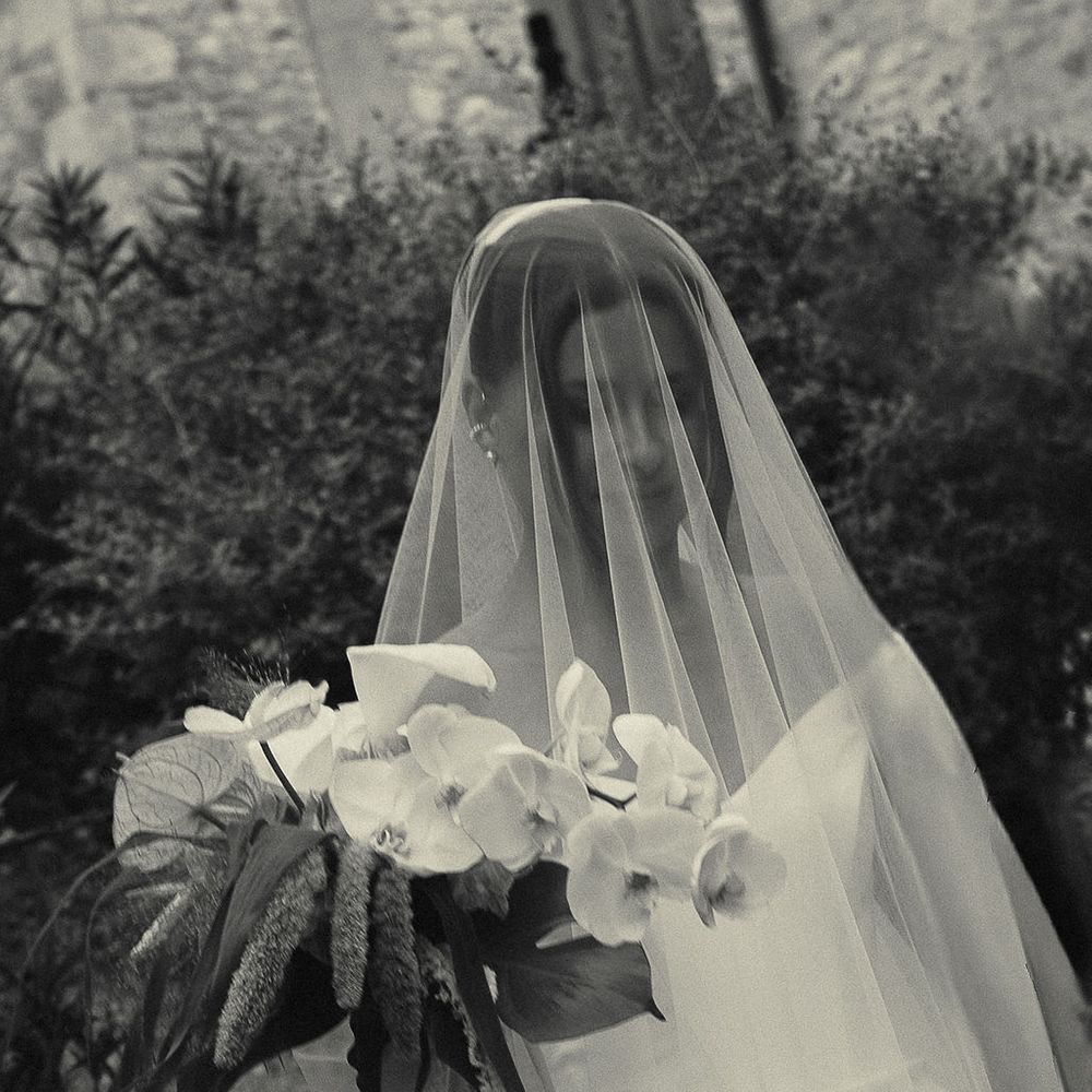 Bride beneath a sheer veil holding an orchid and amaranthus bouquet in a leafy garden