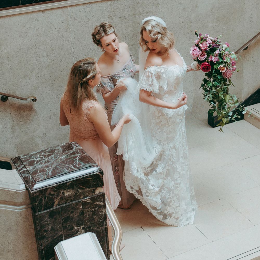 Two bridesmaids in pink adjust the train of a bride's lace gown on a marble staircase