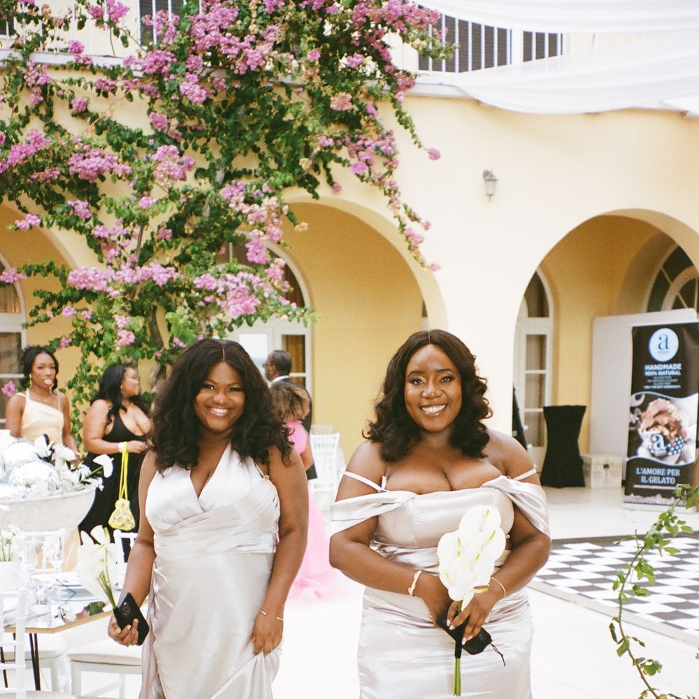 Two smiling bridesmaids in champagne satin gowns beneath blooming bougainvillea in a sunlit courtyard