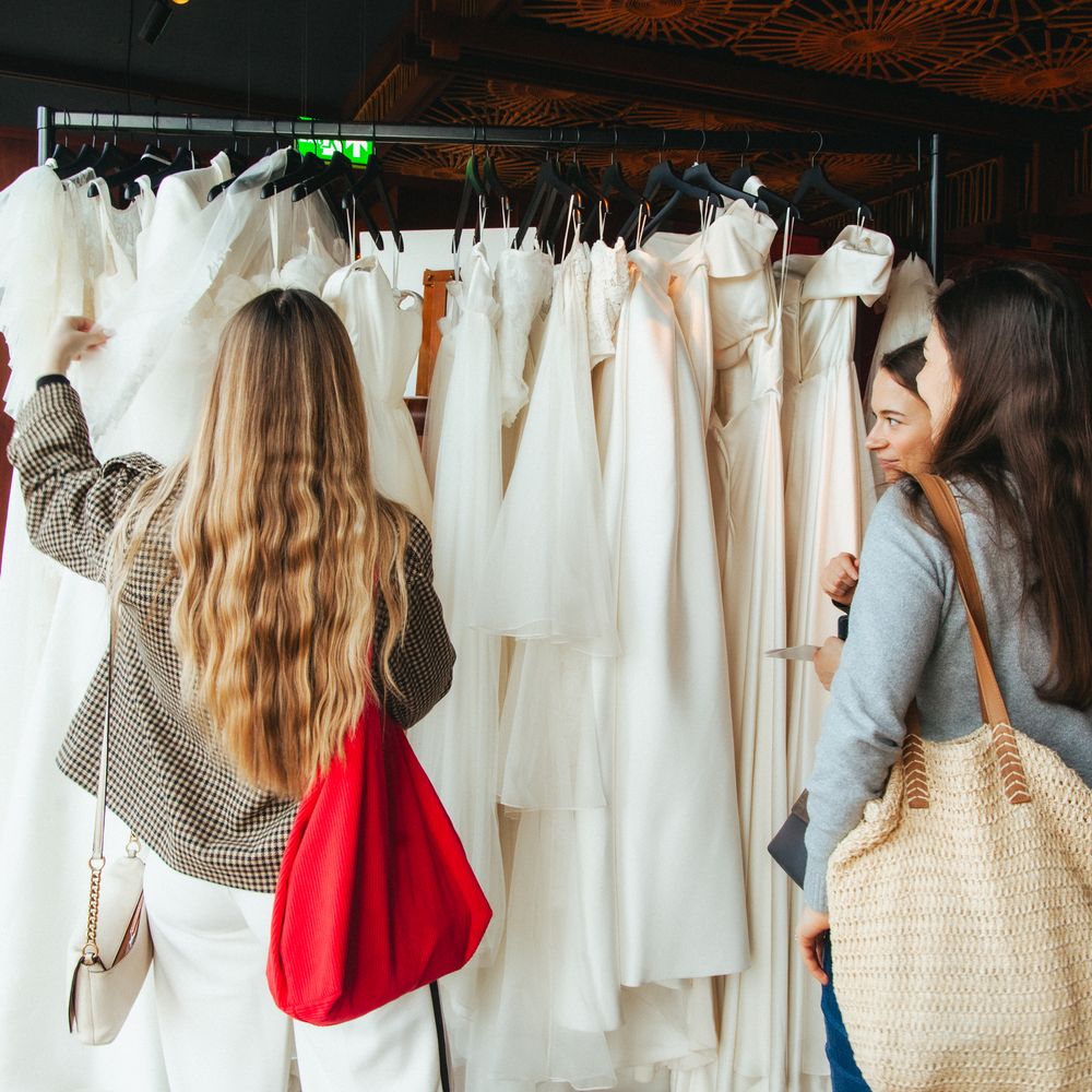 Two women browsing a rail of white wedding dresses in a boutique with an ornate ceiling