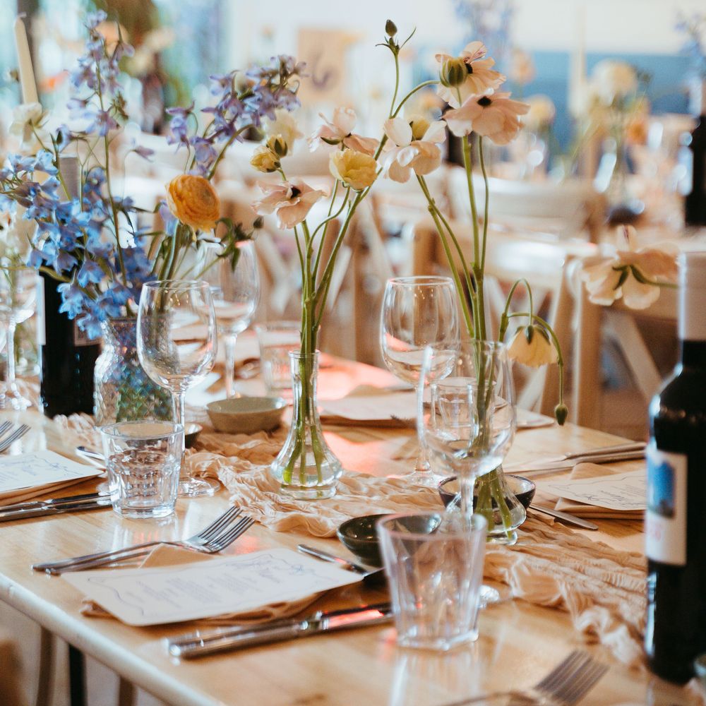 Wooden wedding table with bud vases of blue delphiniums and peach ranunculus beside glassware and menus
