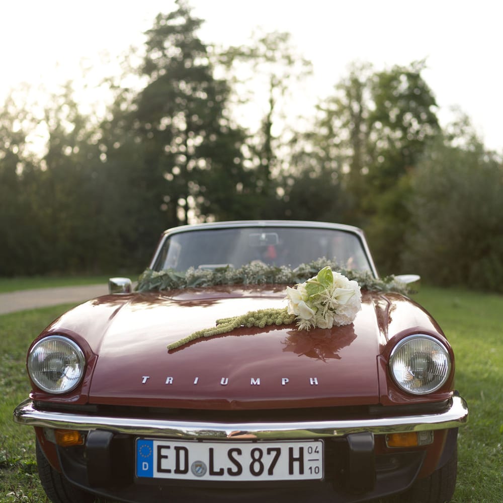 Burgundy vintage Triumph convertible dressed with white florals and foliage on a lawn at dusk