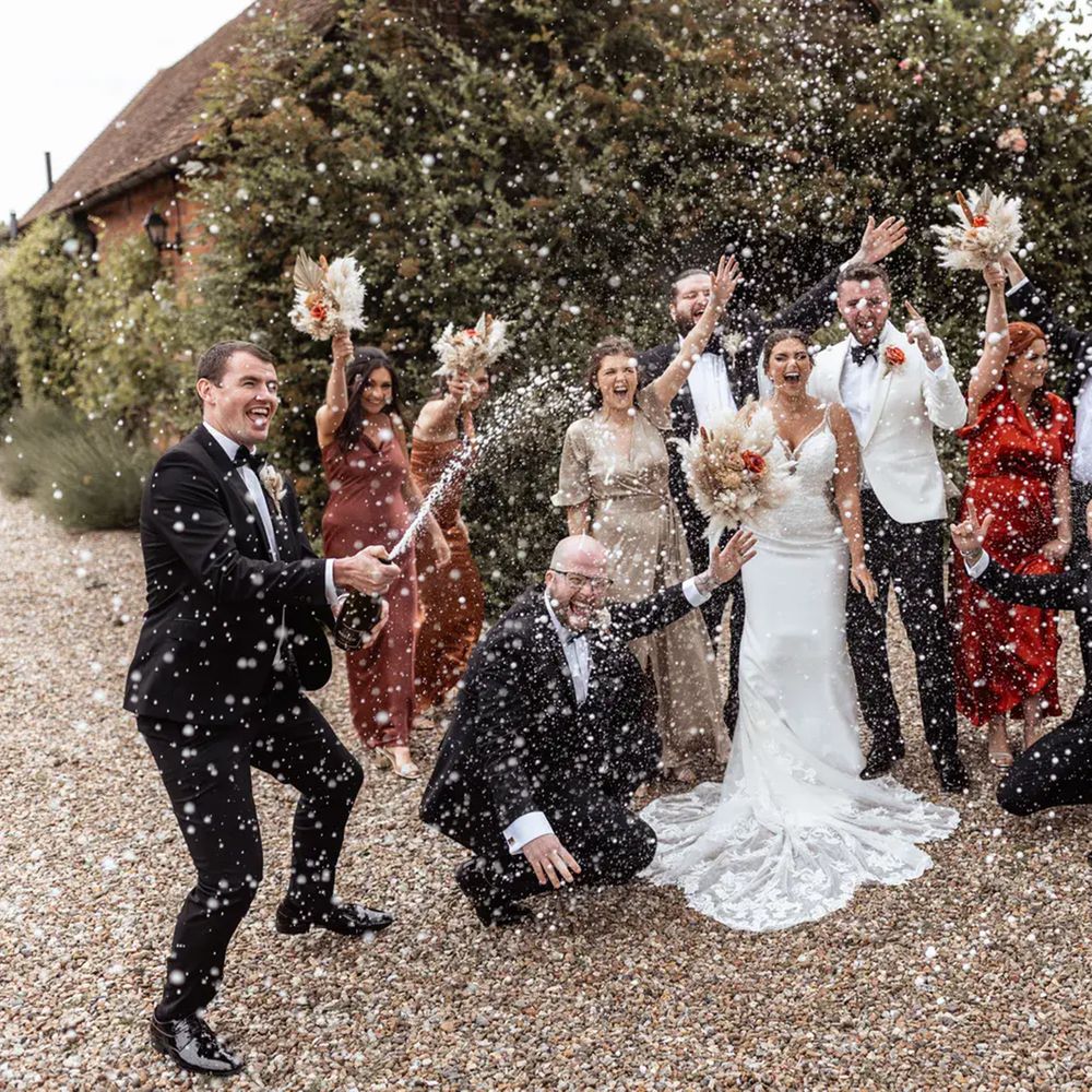Wedding party cheering as champagne sprays over the group outside a brick barn venue