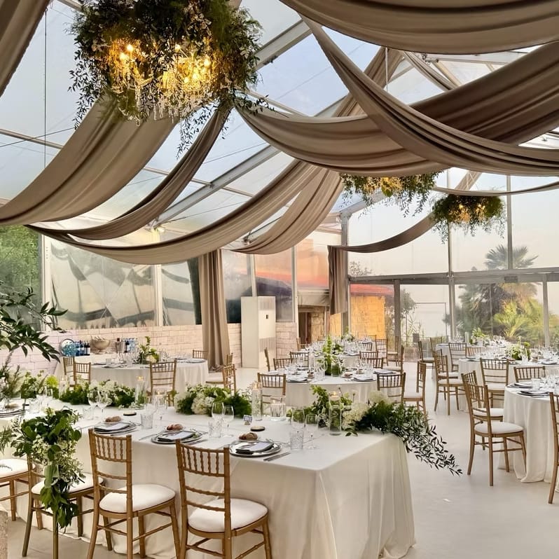 Clear-roofed marquee with draped ceiling, foliage chandeliers and gold chairs set for a wedding breakfast