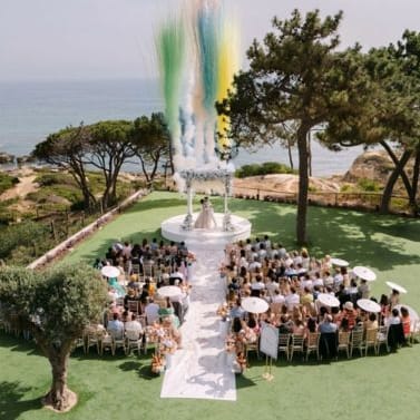 Coastal outdoor wedding ceremony with colourful smoke display above seated guests on green lawn