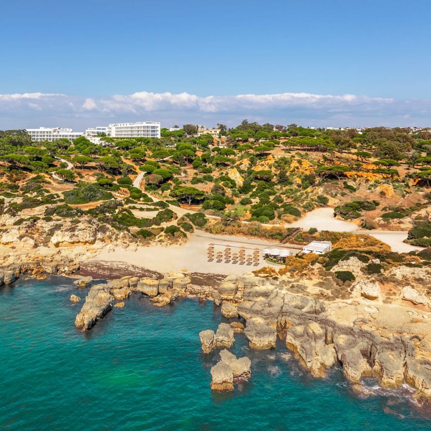Aerial view of a rocky coastline with turquoise sea, sandy cove and clifftop hotel amid pine trees