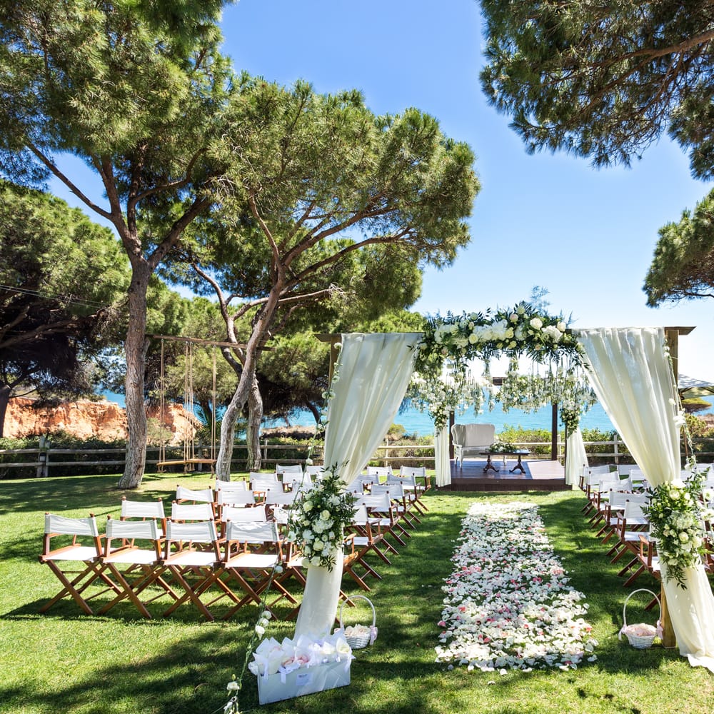 Outdoor coastal ceremony setup with floral arch, petal-strewn aisle and timber chairs beneath pine trees