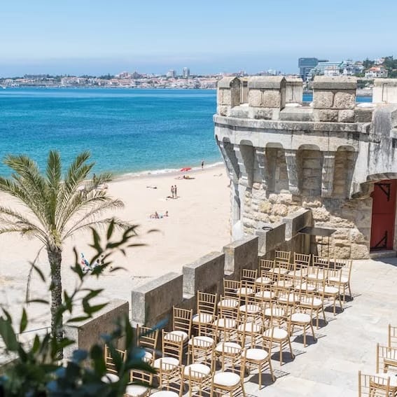 Rows of gold Chiavari chairs set for a ceremony beside a stone fort overlooking a sandy beach and blue sea