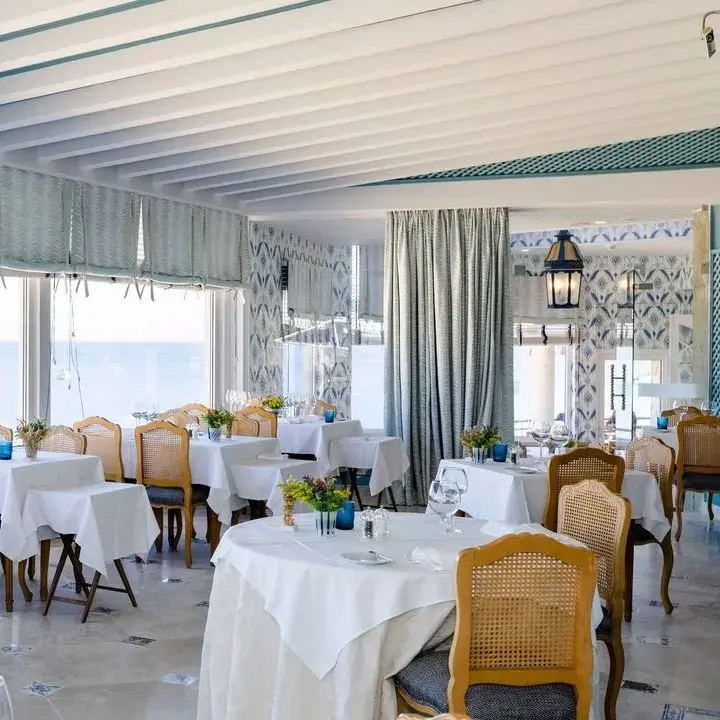 Seaside restaurant with cane dining chairs, white linen tables and floor-to-ceiling ocean views